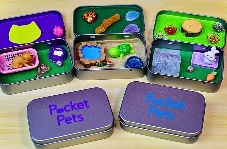 May include: Several small, open metal tins with miniature scenes inside. Each tin contains a different scene, including a cat, a turtle, and a bunny. The lids of the tins are purple and have the words "Pocket Pets" printed on them.
