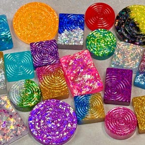 May include: A collection of colorful, glittery resin pieces in various shapes, including circles and rectangles. The pieces feature swirling patterns and a range of colors like blue, gold, pink, and purple. Some have a textured, rippled design.