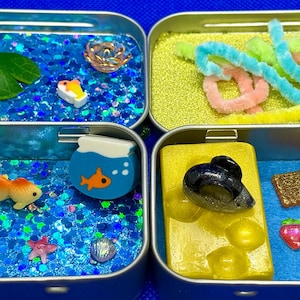 May include: Four small, blue metal tins filled with miniature craft items. The top left tin has a pond theme, the top right has colorful pipe cleaners, the bottom left has an aquarium theme, and the bottom right has a dessert theme.