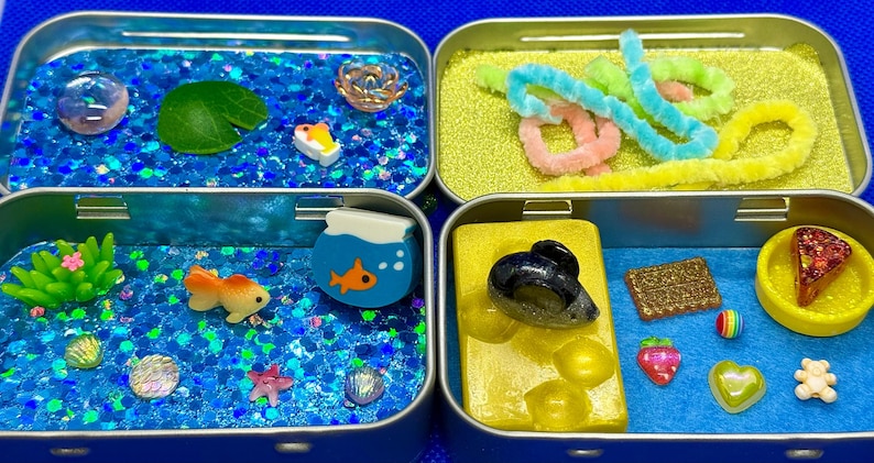 May include: Four small, blue metal tins filled with miniature craft items. The top left tin has a pond theme, the top right has colorful pipe cleaners, the bottom left has an aquarium theme, and the bottom right has a dessert theme.