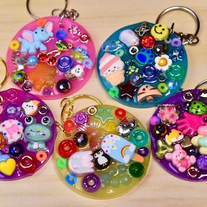 May include: A collection of five colorful resin keychains. Each keychain features a circular design with various embedded charms, including animals, food items, and decorative beads. The keychains have gold-colored key rings.