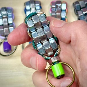 May include: A keychain constructed from silver-colored hexagonal metal nuts woven together with green and white patterned cord. The design features a bright lime green cylindrical bead at the base, attached to a circular gold-toned metal split ring.