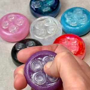 May include: A collection of circular, resin-based sensory fidget tools, each featuring four raised, translucent circular buttons on the surface. The items are available in various colors, including pink, dark blue, light blue, white, black, red, and purple.