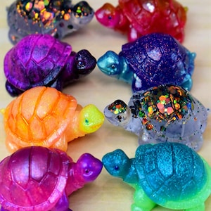 May include: A collection of colorful, glittery turtle figurines in various shades of purple, orange, blue, green, and red. Each turtle has a unique glitter pattern, creating a sparkling effect. The turtles are arranged in a circular pattern.
