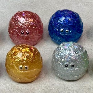 May include: Eight colorful, glittery, round objects with small, white, googly eyes. The colors include green, red, blue, black, purple, gold, silver, and green. The objects appear to be wrapped in a textured material.