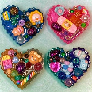 May include: Four heart-shaped resin charms with various embedded embellishments. The charms feature a variety of colors, including blue, pink, gold, and green. Each charm is decorated with miniature objects like a duck, a bear, a popsicle, and a bandage.