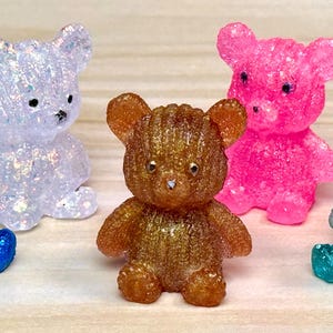 May include: A collection of six glittery teddy bear figurines in various colors: blue, white, brown, pink, and teal. Each bear is sitting with its arms and legs outstretched, showcasing a sparkling, textured surface.