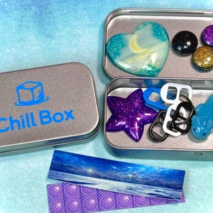 May include: A silver tin box with the words "Chill Box" in blue, containing various small, colorful items. Includes a heart-shaped object, glittery spheres, a star, and a blue shark. Also includes a small landscape image and a purple pop-it toy.