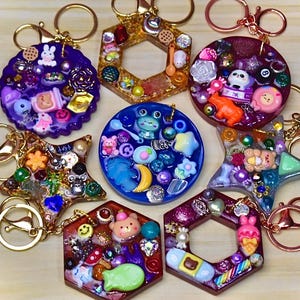 May include: A collection of handmade resin keychains in various shapes, including circles, stars, and hexagons. Each keychain is filled with colorful miniature objects like bears, flowers, and beads. The keychains have gold or rose gold colored key rings.