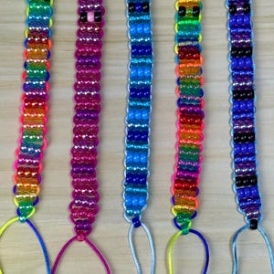 May include: Five beaded keychains in a row. Each keychain features a different color scheme, including rainbow, pink, blue, orange, and blue-purple. The beads are strung on a cord and attached to a metal ring.
