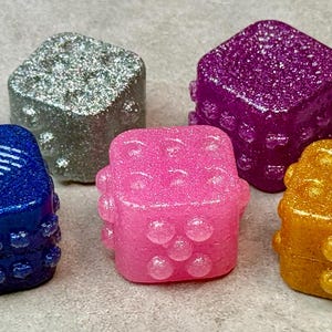 May include: Five colorful, glittery dice in shades of blue, silver, purple, pink, and gold. Each die has a unique color and a textured surface with rounded protrusions. The dice are arranged on a neutral background.