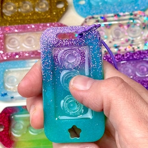 May include: A rectangular resin keychain featuring a purple and teal glitter gradient finish. The keychain has three circular indentations in the center and a small star-shaped cutout at the bottom. A purple cord is attached to the top hole.