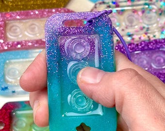 May include: A rectangular resin keychain featuring a purple and teal glitter gradient finish. The keychain has three circular indentations in the center and a small star-shaped cutout at the bottom. A purple cord is attached to the top hole.