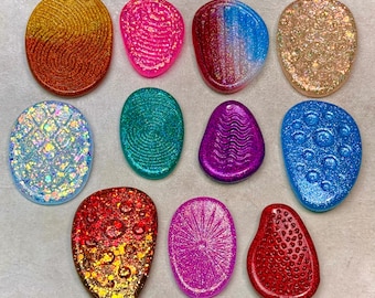 May include: A collection of eleven oval and irregular shaped resin cabochons featuring various textures and finishes. Colors include orange, pink, blue, gold, iridescent white, teal, purple, and red, with glitter, swirl, and dot patterns.