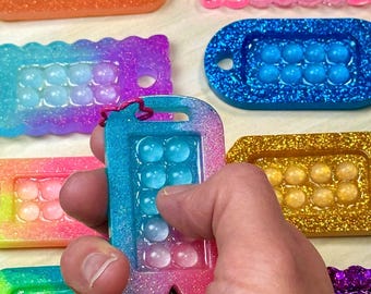 May include: A collection of rectangular resin sensory keychains featuring embedded bubble-like spheres. Each piece displays vibrant glitter finishes in various color gradients, including blue, orange, pink, purple, and gold, with a small hole for attachment.
