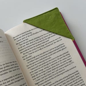 May include: A triangular fabric bookmark with a bright lime green outer layer and a vibrant magenta inner layer, positioned on the top corner of an open paperback book with visible black printed text on white pages.