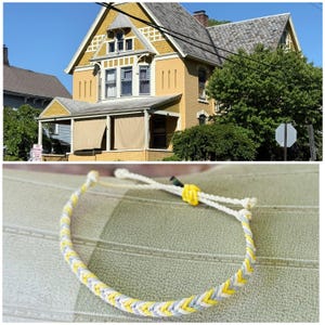May include: A braided bracelet with yellow, gray, and white threads. The bracelet is adjustable with a sliding knot. The background shows a yellow house with a gray roof and green trees.