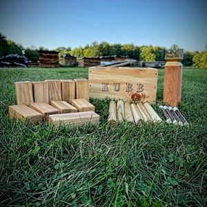 May include: A wooden Kubb game set displayed on a grassy field. The set includes rectangular blocks, wooden dowels, and a wooden box with the word "KUBB" on it. A wooden king piece is also visible. The game is set up outdoors.