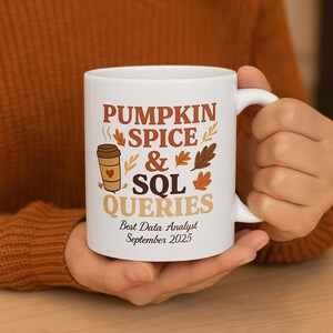 May include: White ceramic mug with the text "PUMPKIN SPICE & SQL QUERIES" in brown and orange. The mug features a coffee cup graphic with a heart and fall leaf accents. The bottom of the mug reads "Best Data Analyst September 2025".