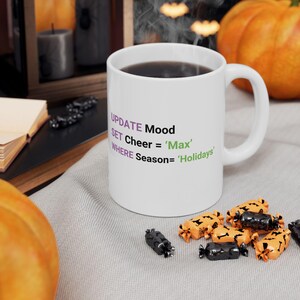 May include: White ceramic mug filled with dark liquid, featuring the text "UPDATE Mood, SET Cheer = 'Max', WHERE Season= 'Holidays'". The mug is surrounded by Halloween-themed candies and pumpkins.