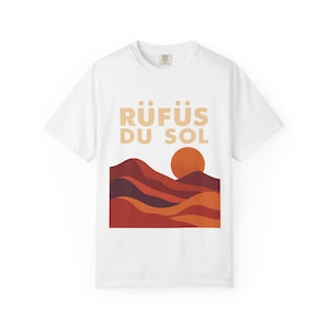May include: White t-shirt featuring the text "RÜFÜS DU SOL" in a light brown font above a graphic of a sunset over a mountain range. The mountain range is depicted in shades of red, orange, and brown.