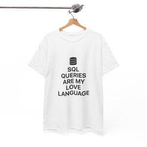 May include: White t-shirt with the text "SQL QUERIES ARE MY LOVE LANGUAGE" in black, and a graphic of three stacked cylinders. The shirt is hanging on a wooden hanger.