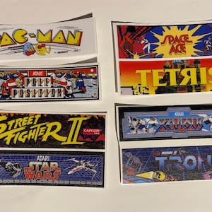 May include: Six vintage arcade game cabinet stickers. The stickers feature the titles Pac-Man, Space Ace, Tetris, Street Fighter II, Star Wars, and Tron. The stickers are printed on white paper with colorful graphics.