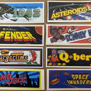 May include: A collection of eight vintage arcade game cabinet artwork panels. The panels are printed on white paper with black, red, blue, yellow, and green colors. The panels feature the names of the games: 1942, Asteroids, Defender, Donkey Kong, BurgerTime, Q*bert, Bomb Jack, and Space Invaders.