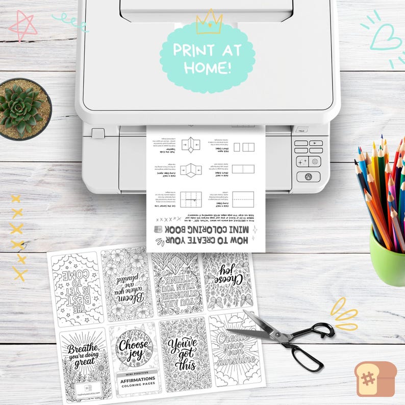 May include: A white printer printing a sheet with instructions for a mini coloring book. Below are affirmation coloring pages with phrases like "Choose Joy" and "You've got this." A small succulent and colored pencils are also visible.