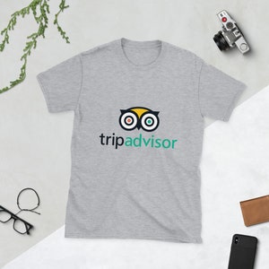May include: A light gray short-sleeve t-shirt featuring the TripAdvisor logo. The logo is an owl-like design with yellow, red, green, and black elements, and the word "tripadvisor" in a teal font. The shirt is made of soft material.