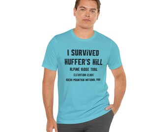 May include: A light blue t-shirt with the text "I Survived Huffer's Hill" in black. Below the text are the words "Alpine Ridge Trail", "Elevation 12,005'", and "Rocky Mountain National Park". The shirt is made of a soft material.