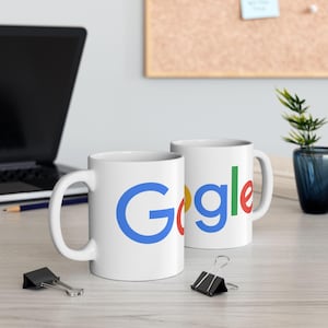 Pode incluir: Duas canecas de cerâmica branca com o logotipo do Google em azul, vermelho, amarelo e verde. As canecas estão sobre uma superfície de madeira clara, com um laptop e material de escritório ao fundo. As canecas têm uma alça curva.