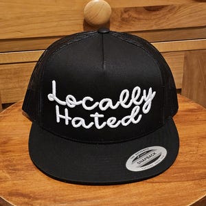 May include: Black and white trucker hat with the text "Locally Hated" in white cursive lettering. The hat has a black mesh back and a black brim with a "The Authentic Yupoong Snapback" label.