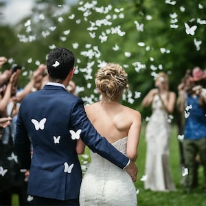 May include: A bride and groom walk down the aisle, surrounded by white butterflies. The bride is wearing a white strapless dress and the groom is wearing a dark blue suit. The couple is smiling and happy.