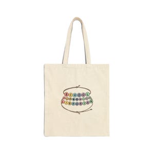 May include: A natural-coloured canvas tote bag featuring a graphic of two brown string bracelets with colourful beads. The top bracelet spells SCHOOL with small icons, and the bottom bracelet spells LIBRARIAN with additional small icons.