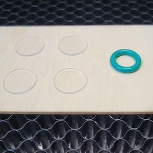 May include: A light-colored wooden board with five clear, circular cutouts and a teal-colored ring. The board is rectangular with rounded corners. The ring is on the right side of the board.