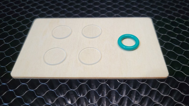 May include: A light-colored wooden board with five clear, circular cutouts and a teal-colored ring. The board is rectangular with rounded corners. The ring is on the right side of the board.