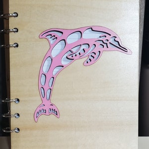 May include: A pink dolphin cutout on a wooden journal with a spiral binding. The dolphin is leaping out of the water.