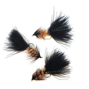 Könnte beinhalten: Vier Angelköder mit schwarzen und braunen Federn und goldenen Haken werden vor einem weißen Hintergrund gezeigt. Die Köder haben ein flauschiges, strukturiertes Aussehen, wobei die schwarzen Federn eine schwanzartige Form bilden. Die braunen Federn sind in der Mitte konzentriert.