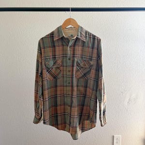 May include: A long-sleeved plaid shirt with a button-down collar. The shirt is green, brown, and orange with a white background. The shirt has two chest pockets.