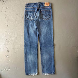 May include: Faded blue denim jeans with a classic five-pocket design. The jeans feature a red logo patch on the back and a worn, distressed look. The jeans have a straight leg cut and a frayed hem.