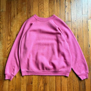 May include: A solid pink sweatshirt with a crew neck and long sleeves. The sweatshirt is made of a soft material and has a ribbed hem and cuffs. The garment is displayed on a wooden surface.