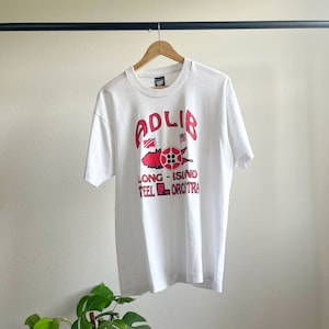 May include: White t-shirt with a red graphic of a boat with the text "Adlib Long Island Steel Orchestra".