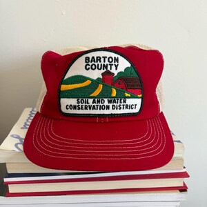 May include: A red and beige trucker hat with a patch that reads "Barton County Soil and Water Conservation District." The patch features a rural landscape with a barn, silo, and rolling hills. The hat has a curved brim and a mesh back.