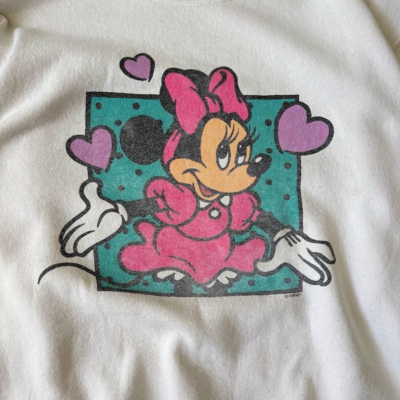 May include: A white sweatshirt featuring a graphic print of Minnie Mouse wearing a pink dress and bow. She is positioned in front of a teal square with black polka dots, surrounded by three purple hearts. The design has a distressed texture.