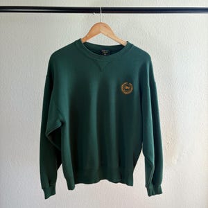 May include: A dark green sweatshirt with a crew neck and long sleeves. The sweatshirt features a gold emblem on the left chest. The garment is hanging on a wooden hanger against a white wall.