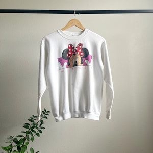 May include: White sweatshirt with a graphic of Minnie Mouse peeking out from behind a pink banner with the word "MINNIE" printed below. The graphic features Minnie Mouse with her signature red polka dot bow.