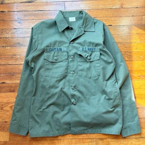 May include: Olive green button-up shirt with two chest pockets. The left pocket has the word "CAPTAIN" embroidered in navy blue, and the right pocket has "U.S. NAVY". The shirt has a classic collar and long sleeves.