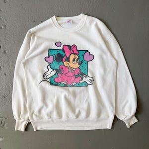 May include: A white crewneck sweatshirt featuring a graphic print of Minnie Mouse wearing a pink dress and bow. The character is centered on a teal polka-dot square background, surrounded by three purple hearts, with long sleeves and ribbed cuffs.
