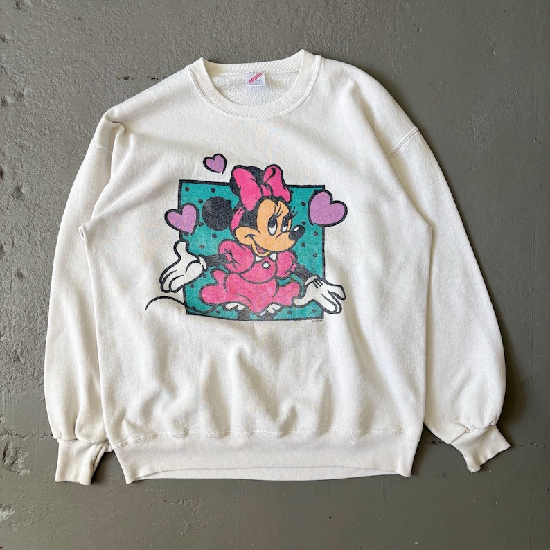 May include: A white crewneck sweatshirt featuring a graphic print of Minnie Mouse wearing a pink dress and bow. The character is centered on a teal polka-dot square background, surrounded by three purple hearts, with long sleeves and ribbed cuffs.